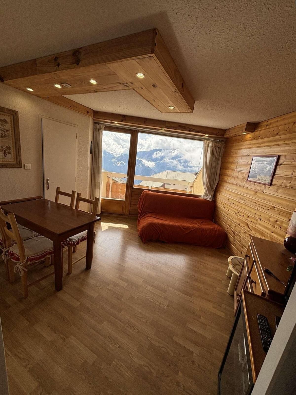 A wooden interior with a dining table, chairs, a sofa, and a view of mountains through a large window.