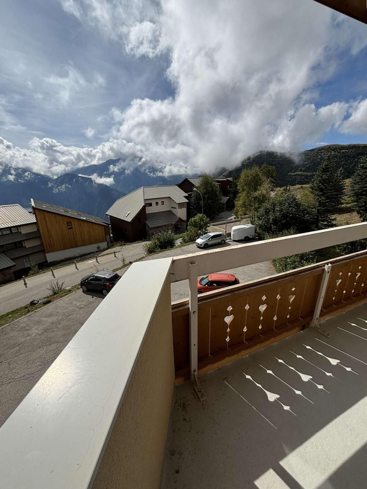 Un balcon offrant une vue sur une chaîne de montagnes, un parking avec des voitures et des bâtiments à l’architecture traditionnelle.