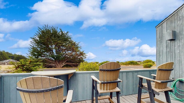 A deck with wooden Adirondack chairs and a table, overlooking a blue-painted wall and a tree. - Sea Ranch 2BR home with private hot tub & game room — dog friendly (Sea Ranch)