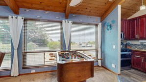 A room with a foosball table, large windows with curtains, a kitchen area, and a blue accent wall. - Sea Ranch 2BR home with private hot tub & game room — dog friendly (Sea Ranch)