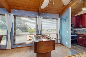 A room with a foosball table, large windows with curtains, a kitchen area, and a blue accent wall.