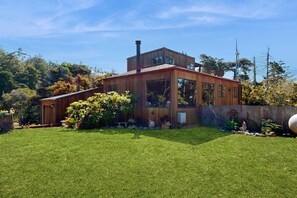 A modern wooden house with a large glass window, a chimney, and a well-maintained lawn. - Gualala 1BR + loft home with partial ocean views — walk to town (Gualala)