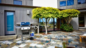 A modern outdoor area with a barbecue grill, a stone-tiled patio, and a view of a building with large windows. - Coastal Luxury: Chef Kitchen, Ocean Views, Hot Tub (Sea Ranch)