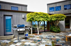A modern outdoor area with a barbecue grill, a stone-tiled patio, and a view of a building with large windows. - Coastal Luxury: Chef Kitchen, Ocean Views, Hot Tub (Sea Ranch)