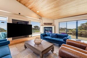 A modern living room with a flat-screen TV, a blue sofa, a wooden coffee table, and a view of the outdoors through large windows. - Sunsets over the ocean, waves, whales, serenity, luxury on the bluff (Sea Ranch)