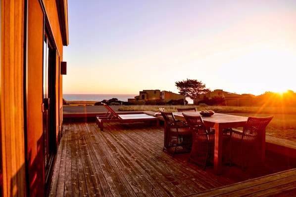 A wooden deck with outdoor furniture, overlooking a beach and a distant hill. - Coastal Luxury: Chef Kitchen, Ocean Views, Hot Tub (Sea Ranch)