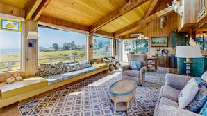 Un interior de madera con un ventanal grande, un sofá, una silla, una mesa de comedor y una zona de cocina.