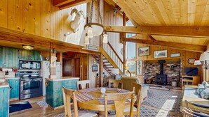 Interior de una cabaña de madera con cocina, comedor y sala de estar, que incluye una chimenea y una escalera.
