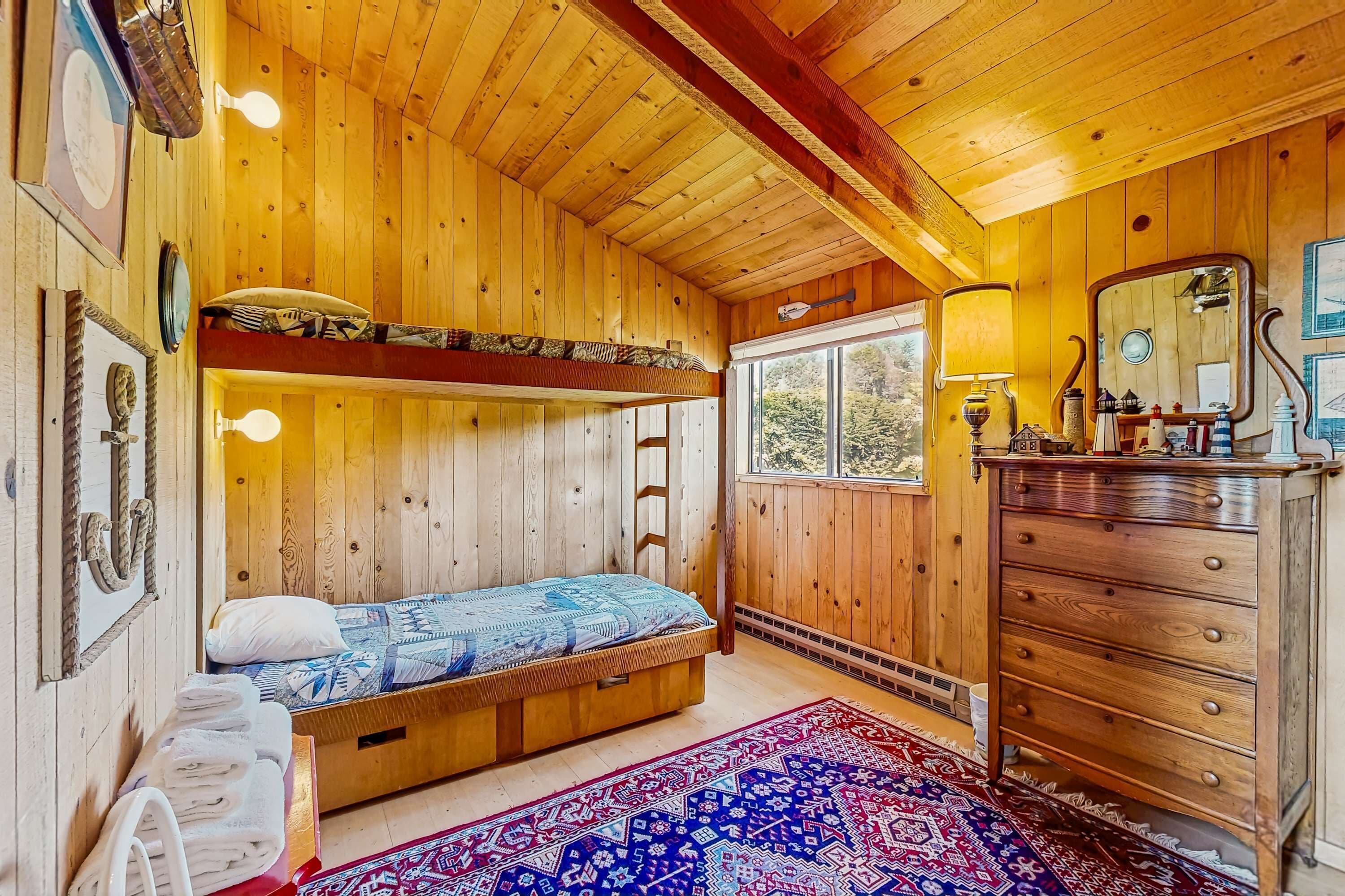 A wooden cabin room with a bunk bed, a dresser, a mirror, and a window with a view of trees.