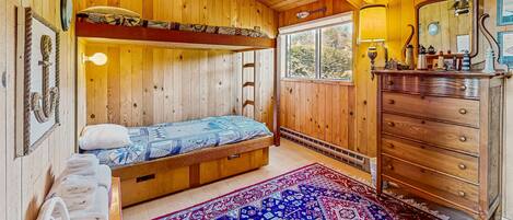 A wooden cabin room with a bunk bed, a dresser, a mirror, and a window with a view of trees.