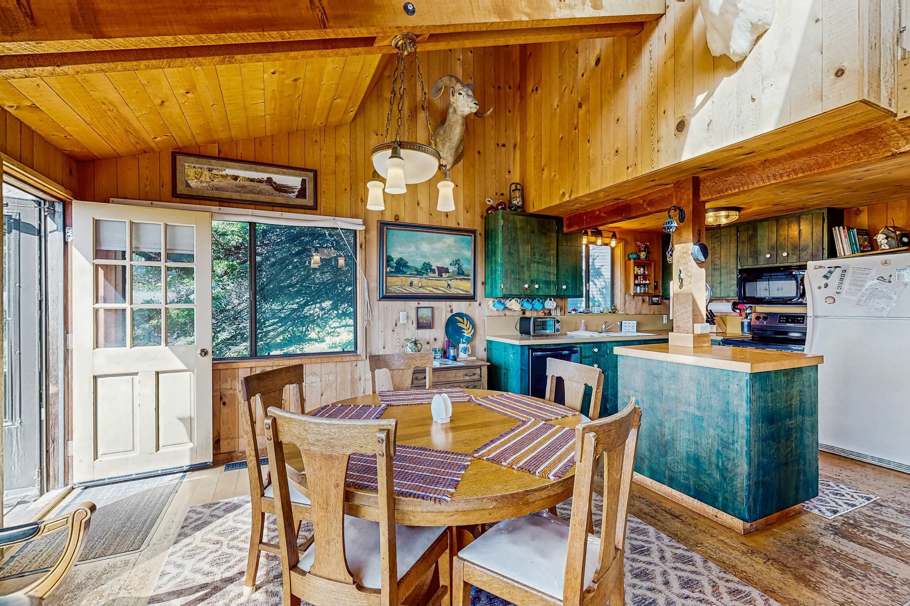 A wooden cabin interior with a dining table, chairs, a kitchen area, and a mounted animal head on the ceiling.
