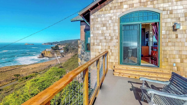 Un bâtiment en bois doté d’un balcon donnant sur l’océan, d’un mur en pierre et d’une terrasse en bois avec un banc.