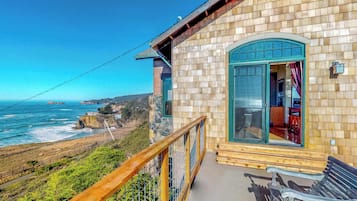 A wooden building with a balcony overlooking the ocean, a stone wall, and a wooden deck with a bench.