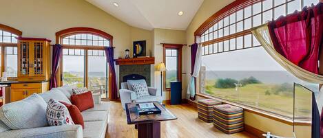 A spacious living room with a sofa, a coffee table, and a fireplace. Large windows offer a view of the outdoors.