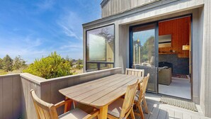 A wooden deck with a table and chairs, a sliding glass door to a room with a fireplace, and a view of the outdoors. - Sea Ranch 2BR ocean-view home with private hot tub & courtyard (Sea Ranch)