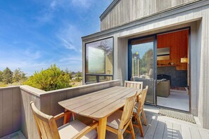 A wooden deck with a table and chairs, a sliding glass door to a room with a fireplace, and a view of the outdoors. - Sea Ranch 2BR ocean-view home with private hot tub & courtyard (Sea Ranch)