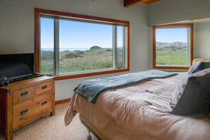 Ein Schlafzimmer mit einem Bett, einer Holzkommode, einem Fernseher und zwei Fenstern mit Blick auf eine grüne Landschaft.