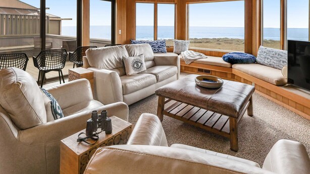 Amplia sala de estar con un gran ventanal con vista a la playa, un sofá de cuero, una mesa de centro y un televisor.