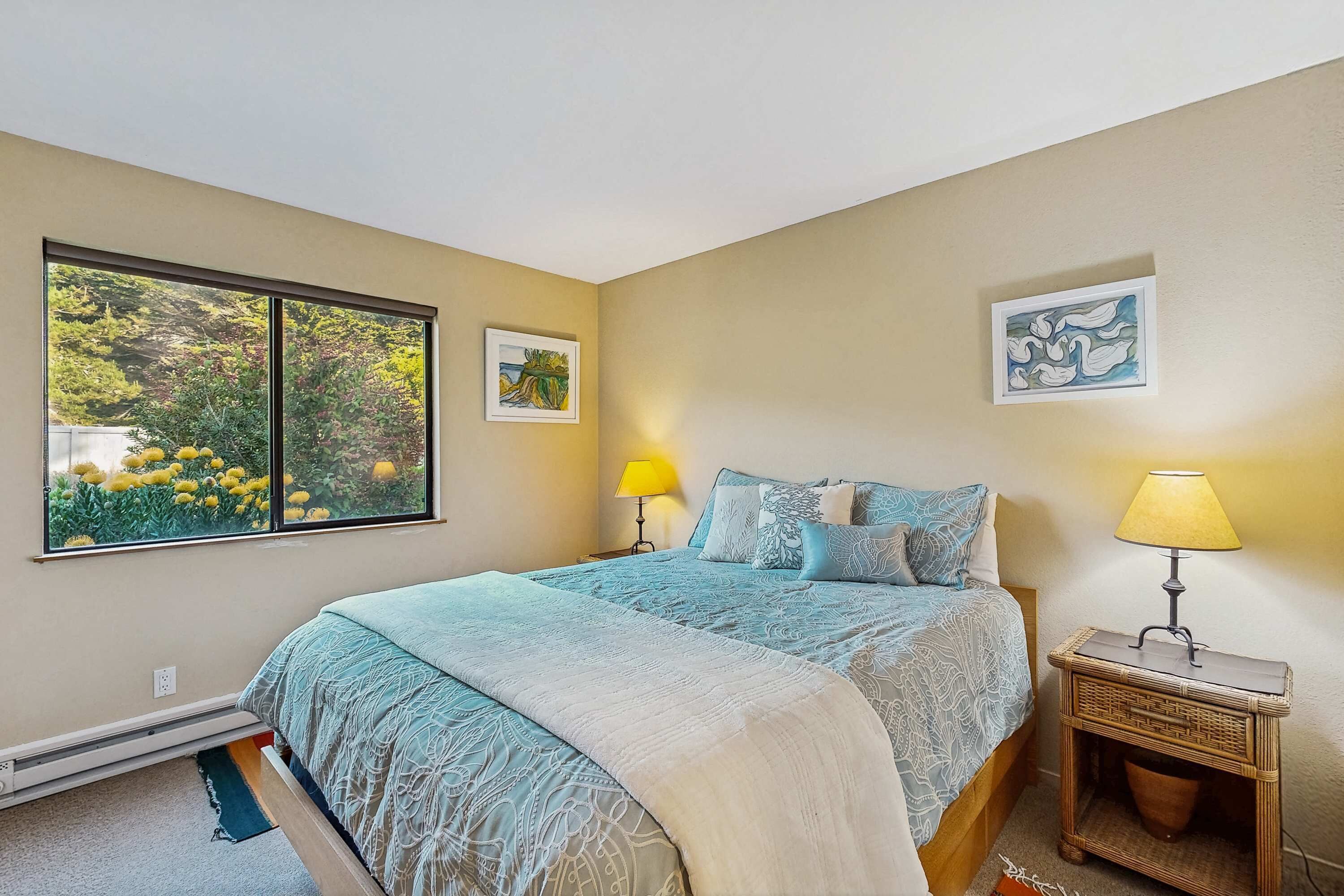 A bedroom with a large window, a bed with patterned bedding, two bedside lamps, and a wall-mounted picture.