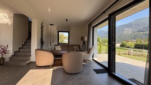 A modern living room with a sofa, ottoman, and a dining table. There is a staircase leading to another level, and a large sliding glass door offering a view of a mountainous landscape. - New luxury villa—for outdoor adventures in both summer and winter! (Gundersheim)