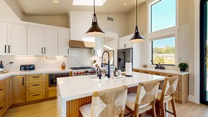 A modern kitchen with white cabinets, a large island with bar stools, pendant lighting, and a view through a window. - Luxe newly built Sea Ranch home with private hot tub & ocean views (Sea Ranch)