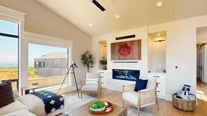 A modern living room with a large window, a coffee table with a plant, a TV, and a cozy seating area. - Luxe newly built Sea Ranch home with private hot tub & ocean views (Sea Ranch)
