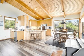 A modern kitchen with wooden cabinets, a central island with stools, a dining area with chairs, and large windows offering a view of the outdoors. - Meadow home with private hot tub on the golf course (Sea Ranch)