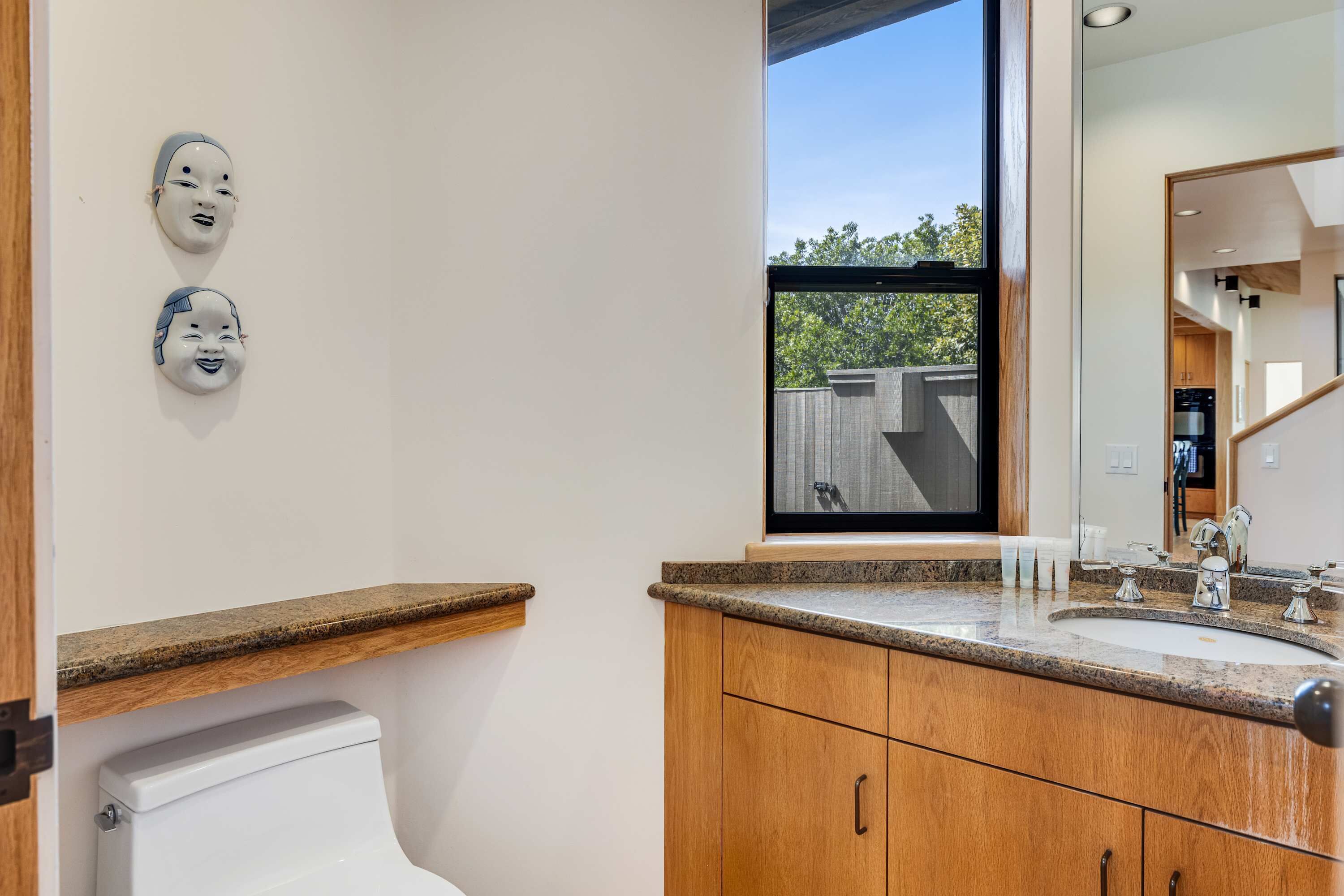 Un baño con inodoro, armarios de madera, encimera de granito, lavabo y ventana con vista a árboles.