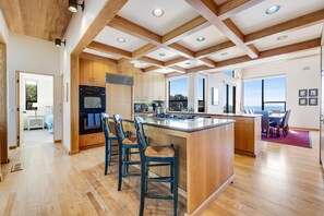 A spacious kitchen with wooden beams, a large island, bar stools, and a view through the windows. - Sea Ranch 4BR architectural home with ocean views & private hot tub (Sea Ranch)