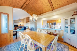 A wooden dining table with chairs, a chandelier, and a kitchen area with cabinets and a microwave. - Ocean view home with fireplace, deck, private hot tub & grill (Sea Ranch)