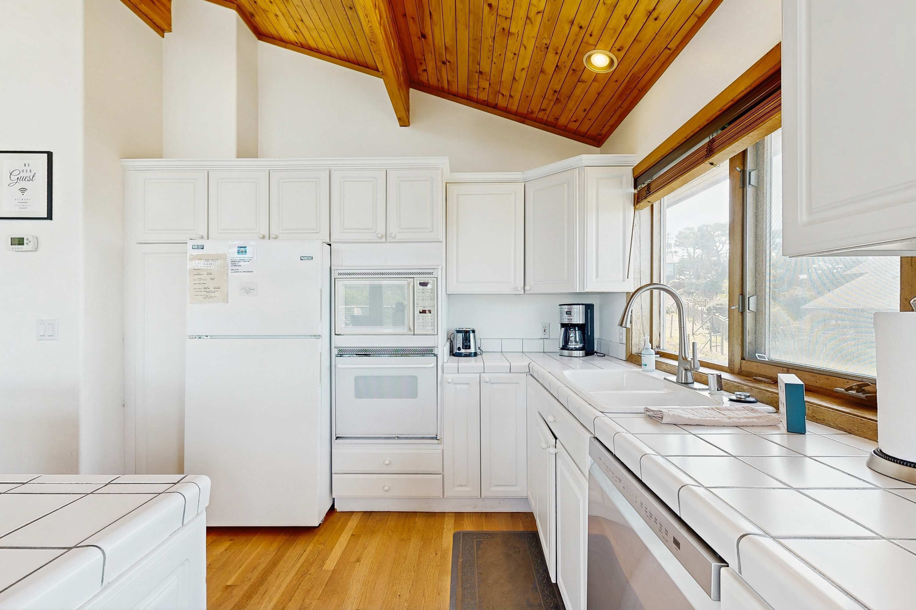 A kitchen with white cabinets, a refrigerator, an oven, a microwave, a coffee maker, and a window with a view.