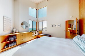 A bedroom with a large bed, a wooden dresser, a seating area with a bench, a round mirror, and a view of the outdoors. - Oceanfront home near Shell Beach & Bluff Trail with deck & ocean views (Sea Ranch)
