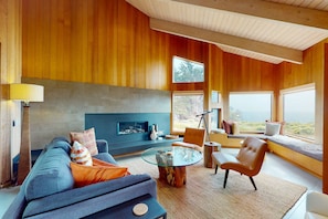 A modern living room with a fireplace, a sofa, a coffee table, and a view through large windows. - Oceanfront home near Shell Beach & Bluff Trail with deck & ocean views (Sea Ranch)