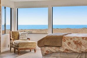 A bedroom with a large window overlooking the beach, a chair with a cushion, and a bed with a patterned bedspread. - Dog-friendly home with cove views, a private hot tub, & rec center access (Sea Ranch)