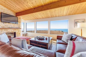 A modern interior with a wooden ceiling, a flat-screen TV, a stone wall, a leather sofa, a dining table, and a view of the beach. - Dog-friendly home with cove views, a private hot tub, & rec center access (Sea Ranch)