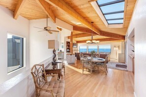 A spacious room with a high ceiling, wooden beams, a skylight, a dining area with a table and chairs, and a view of the ocean. - Dog-friendly home with cove views, a private hot tub, & rec center access (Sea Ranch)