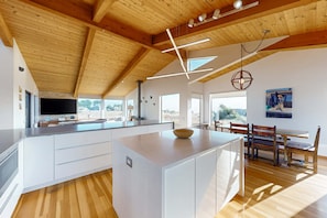 A modern kitchen with wooden beams, a large island, and a dining area with a bench. - Ocean-view meadow home with private hot tub, dogs welcome (Sea Ranch)