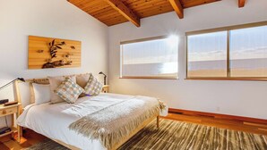 Quarto com cama, cabeceira de madeira, mesas de cabeceira, criadeira e uma obra de arte pendurada na parede, além de uma janela grande com vista.