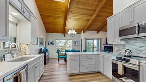 A modern kitchen with wooden beams, a central island, stainless steel appliances, and a dining area with blue chairs. - Gualala 3BR updated coastal home with ocean views & private deck (Gualala)