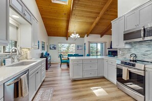 A modern kitchen with wooden beams, a central island, stainless steel appliances, and a dining area with blue chairs. - Updated coastal home with tranquil ocean views, gas grill, private deck (Gualala)