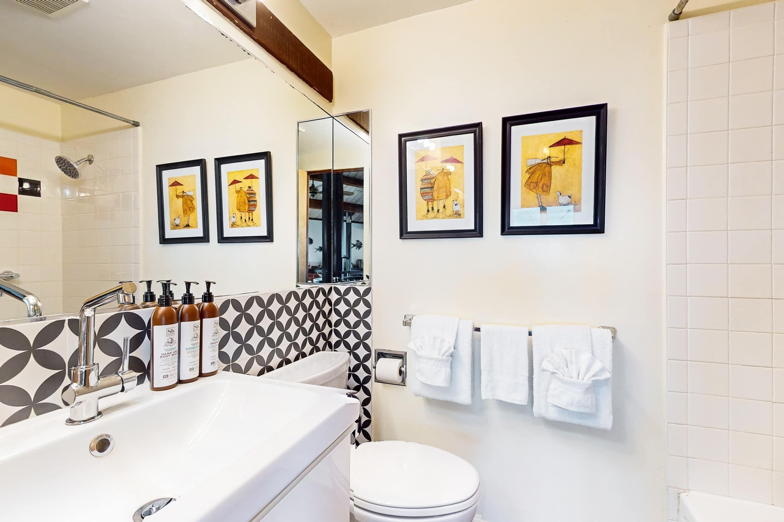 A bathroom with a white sink, a toilet, a shower, and framed artwork on the wall.