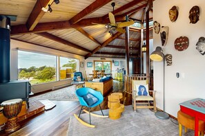 A spacious living area with a high ceiling, wooden beams, a blue rocking chair, a wooden coffee table, a fireplace, and a view of the outdoors. - Newly remodeled hexagon home with hot tub, dogs welcome (Sea Ranch)