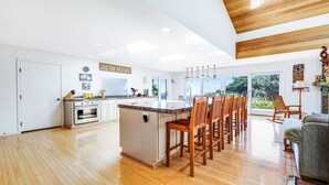 A spacious kitchen with a central island, wooden flooring, and large windows offering a view of greenery. - Gualala 3BR ocean-view home with hot tub — walk to Cooks Beach (Gualala)