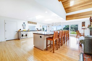 A spacious kitchen with a central island, wooden flooring, and large windows offering a view of greenery. - Gualala 3BR ocean-view home with hot tub — walk to Cooks Beach (Gualala)
