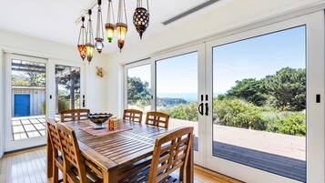 A dining area with a wooden table, chairs, hanging lanterns, and large windows offering a view of greenery and a distant landscape.