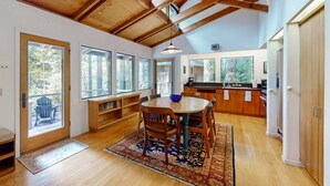 A spacious kitchen with wooden beams, a dining area with a round table and chairs, and a view through large windows. - Sea Ranch 2BR forested home with deck & grill near the Lodge — dog friendly (Sea Ranch)