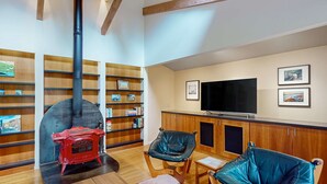 A cozy living room with a red wood-burning stove, wooden shelves, a television, and two blue chairs. - Sea Ranch 2BR forested home with deck & grill near the Lodge — dog friendly (Sea Ranch)