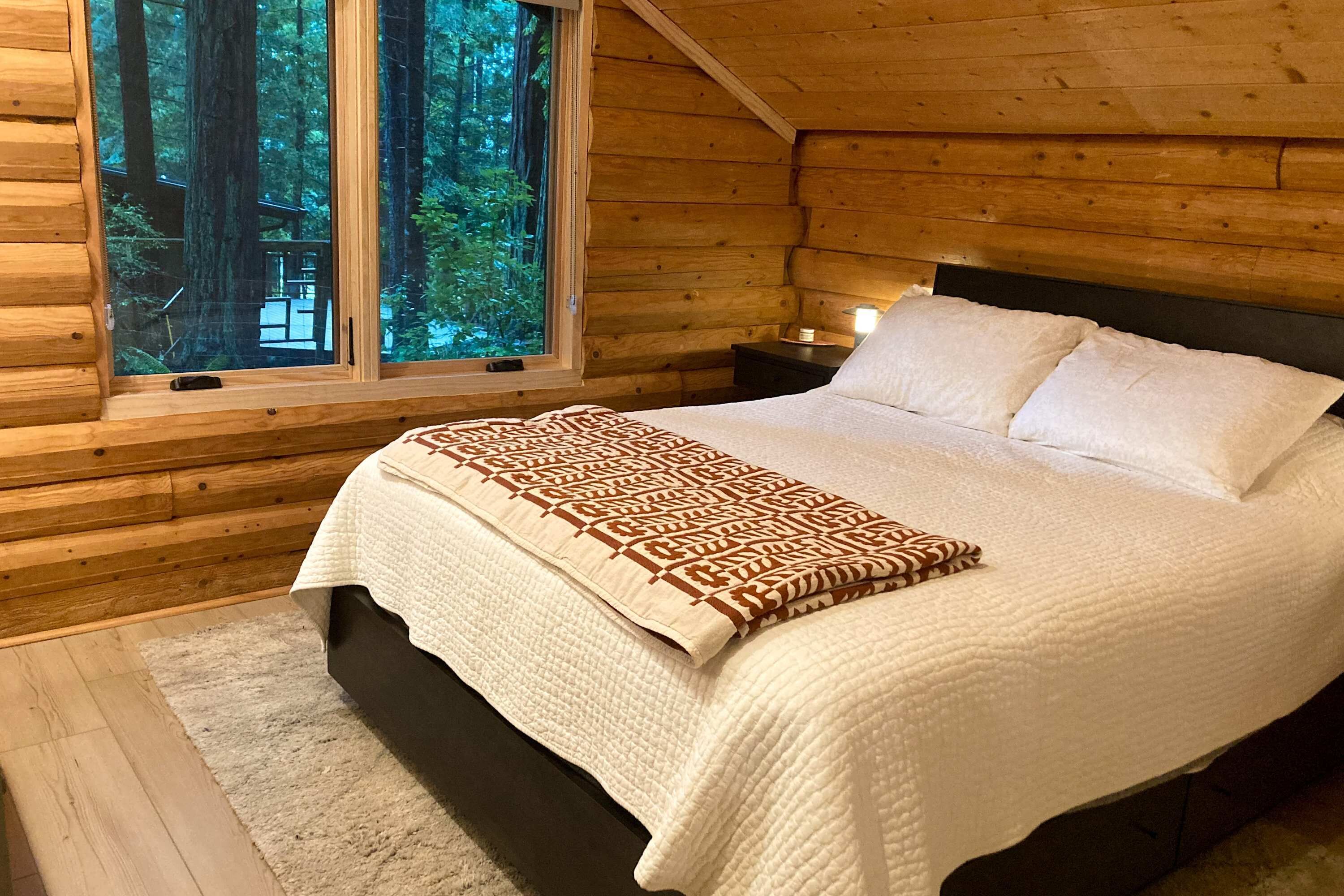 A wooden cabin bedroom with a bed, a nightstand, a window with a view of trees, and a blanket.