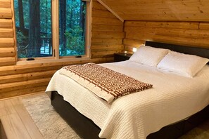 A wooden cabin bedroom with a bed, a nightstand, a window with a view of trees, and a blanket. - Gualala 2BR log cabin in the redwoods — fire-table deck & EV charger (Gualala)