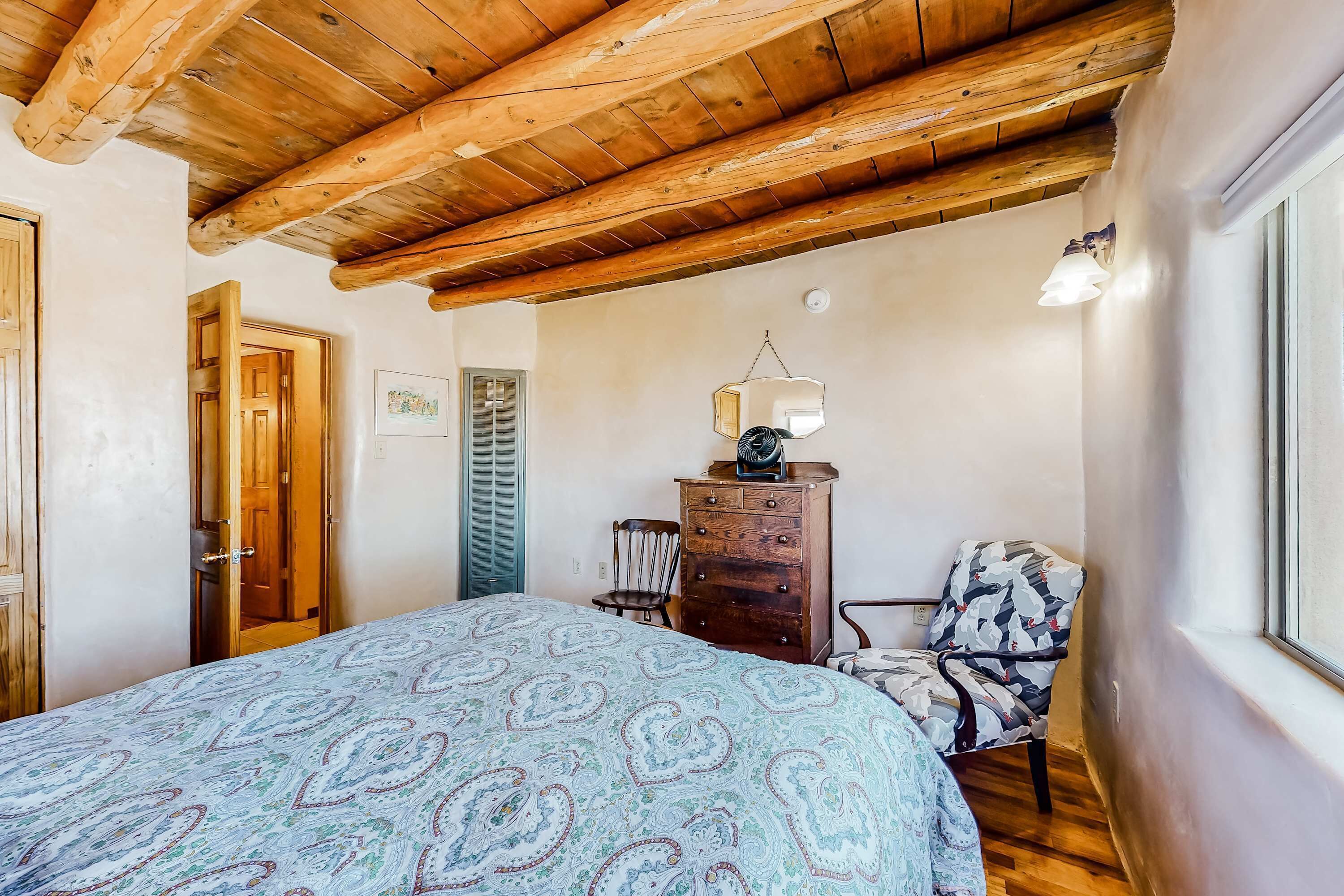 A bedroom with a wooden ceiling, a bed with a patterned bedspread, a dresser, a chair, and a window.